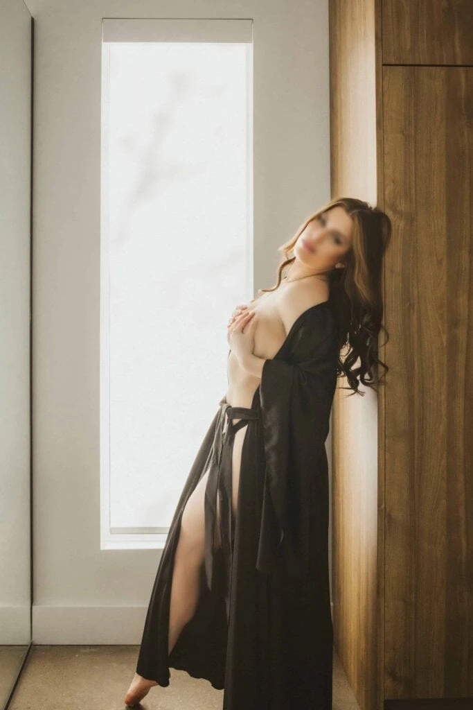 Ava Marie, dressed in a flowing black robe, stands gracefully by a frosted glass window. The soft light casts a warm glow that emphasises her thought-provoking elegance. Ava Marie Halifax's Elite Independent Companion
